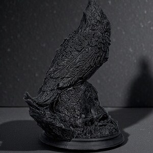 May include: A black raven sculpture with detailed feather and texture, perched on a textured base. The raven is sculpted in a realistic style, with a slightly raised head. The sculpture is set against a dark background.