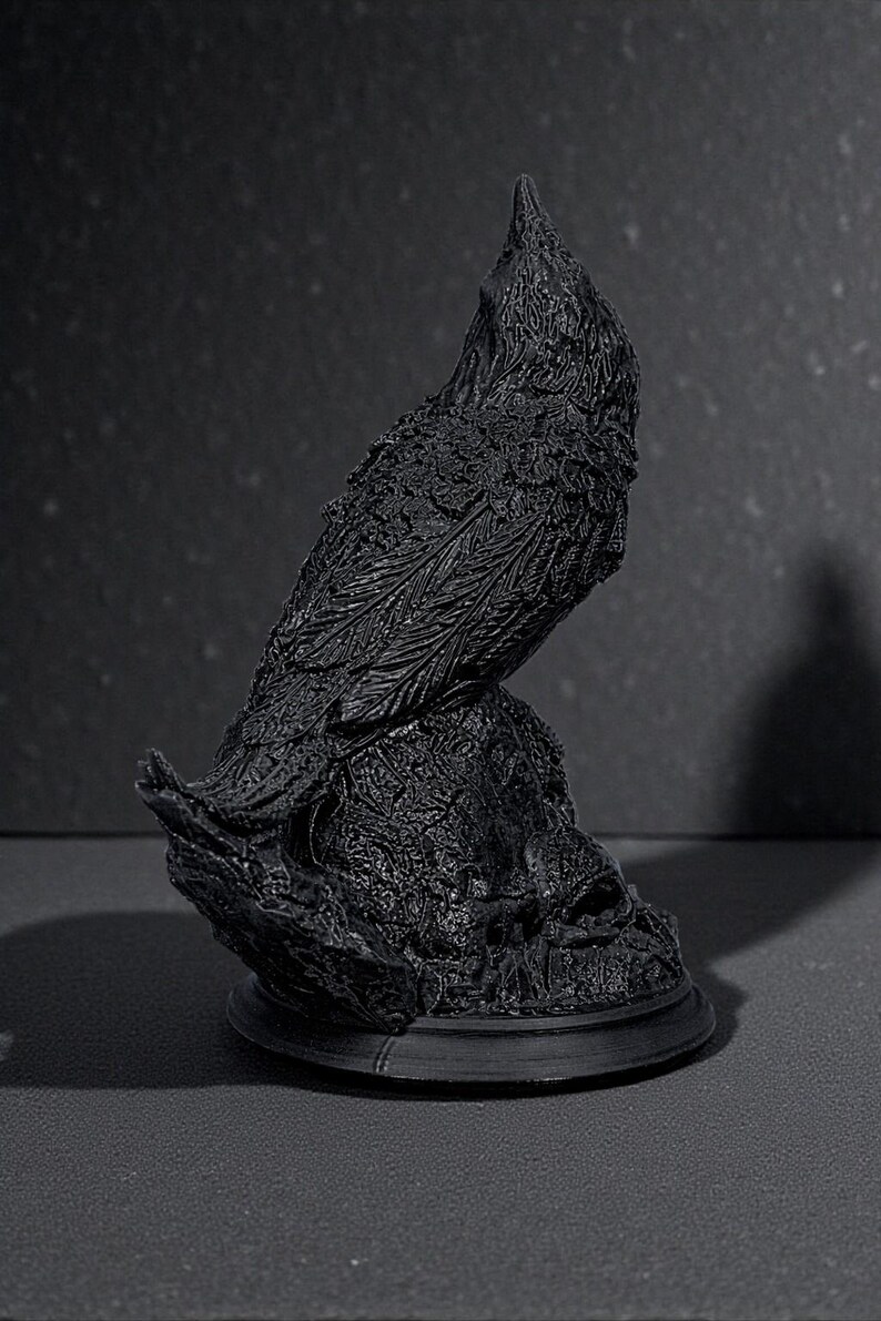 May include: A black raven sculpture with detailed feather and texture, perched on a textured base. The raven is sculpted in a realistic style, with a slightly raised head. The sculpture is set against a dark background.
