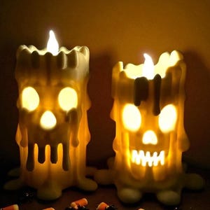 May include: Two Halloween-themed candle decorations. The off-white candles have a melted wax appearance and are shaped like skulls with glowing eyes and mouths. The candles have a flickering flame and are surrounded by candy corn and plastic spiders.
