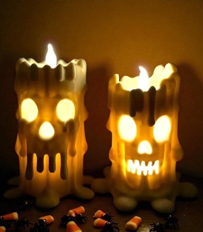 May include: Two Halloween-themed candle decorations. The off-white candles have a melted wax appearance and are shaped like skulls with glowing eyes and mouths. The candles have a flickering flame and are surrounded by candy corn and plastic spiders.