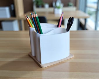 Op de afbeelding: Een witte bureau-organizer met drie compartimenten, met kleurpotloden en een zwarte schaar. De organizer staat op een licht houten basis, geplaatst op een houten tafel. De potloden zijn verschillende kleuren.