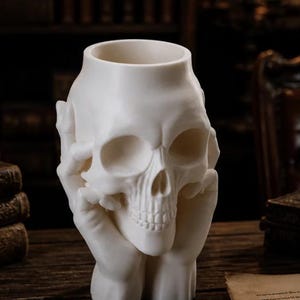 May include: A white skull-shaped decorative vase. The skull is held by two hands, with the fingers gently touching the skull's temples. The vase has a hollow interior, perfect for holding small items or as a unique decorative piece.