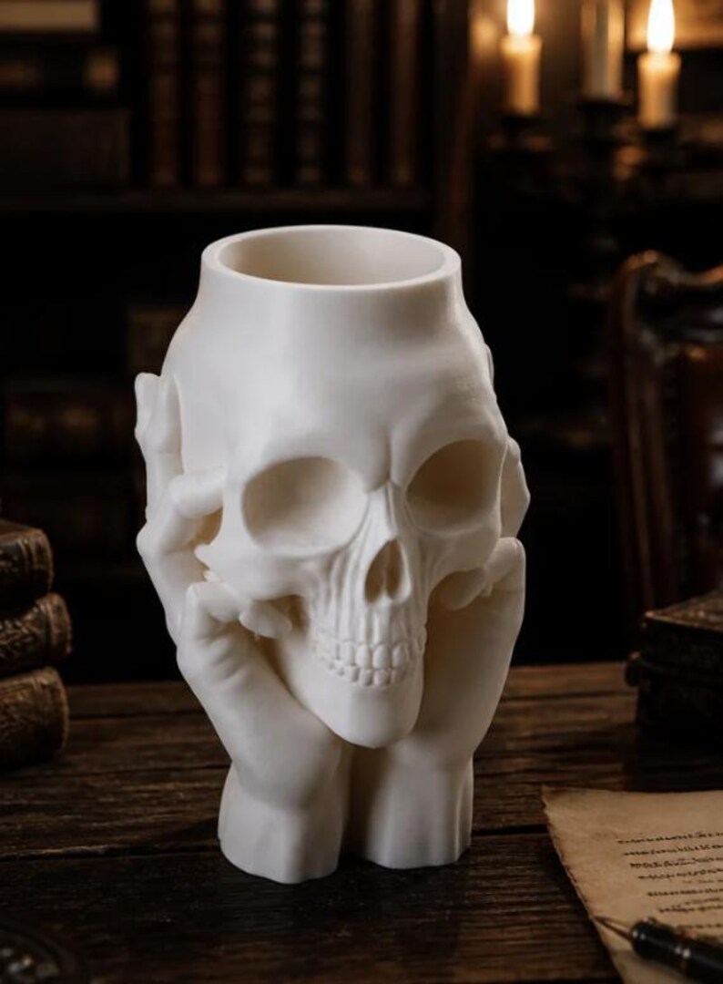 May include: A white skull-shaped decorative vase. The skull is held by two hands, with the fingers gently touching the skull's temples. The vase has a hollow interior, perfect for holding small items or as a unique decorative piece.