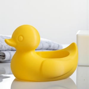 Op de afbeelding: Een heldergele, eendvormige container. De container heeft een afgerond lichaam met een kleine opening aan de bovenkant. De eend heeft een klein, rond oog en een gebogen snavel. De container staat op een wit oppervlak.