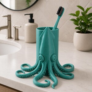 May include: A teal octopus-shaped toothbrush holder with a toothbrush inside. The holder has a cylindrical body with an octopus design, including tentacles and large eyes. The toothbrush has a teal handle and black bristles. The holder sits on a white countertop.