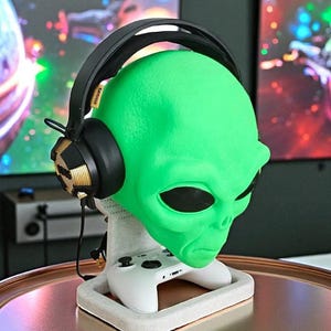May include: A green alien head-shaped headphone stand with a white base holding a white game controller. Black and gold headphones rest on the alien head. The stand is on a copper-colored surface.