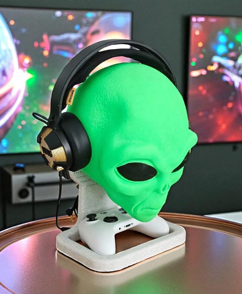 May include: A green alien head-shaped headphone stand with a white base holding a white game controller. Black and gold headphones rest on the alien head. The stand is on a copper-colored surface.