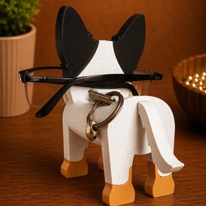 May include: A decorative dog-shaped eyeglasses holder. The holder is white with black and speckled accents, featuring a key ring and eyeglasses. The dog's ears are black, and the feet are orange. The eyeglasses are black-framed.