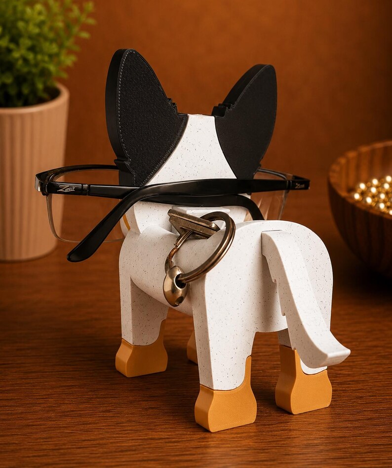 May include: A decorative dog-shaped eyeglasses holder. The holder is white with black and speckled accents, featuring a key ring and eyeglasses. The dog's ears are black, and the feet are orange. The eyeglasses are black-framed.