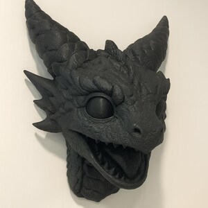 May include: A black dragon head sculpture with textured details, including horns, scales, and an open mouth revealing teeth. The dragon head is mounted on a white wall, showcasing a detailed fantasy art piece.