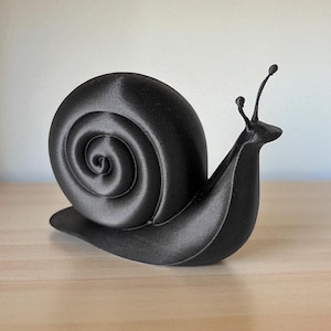 May include: A black 3D-printed snail ornament. The snail features a spiralled shell and two antennae. The ornament is placed on a light-coloured wooden surface.