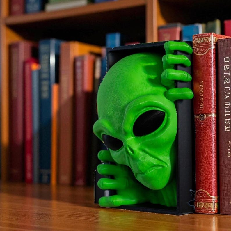 May include: A vibrant green alien head bookend with large black eyes and a furrowed brow. The alien is peeking out from a black rectangular frame, with its hands gripping the sides. The bookend is positioned amongst a collection of books on a shelf.