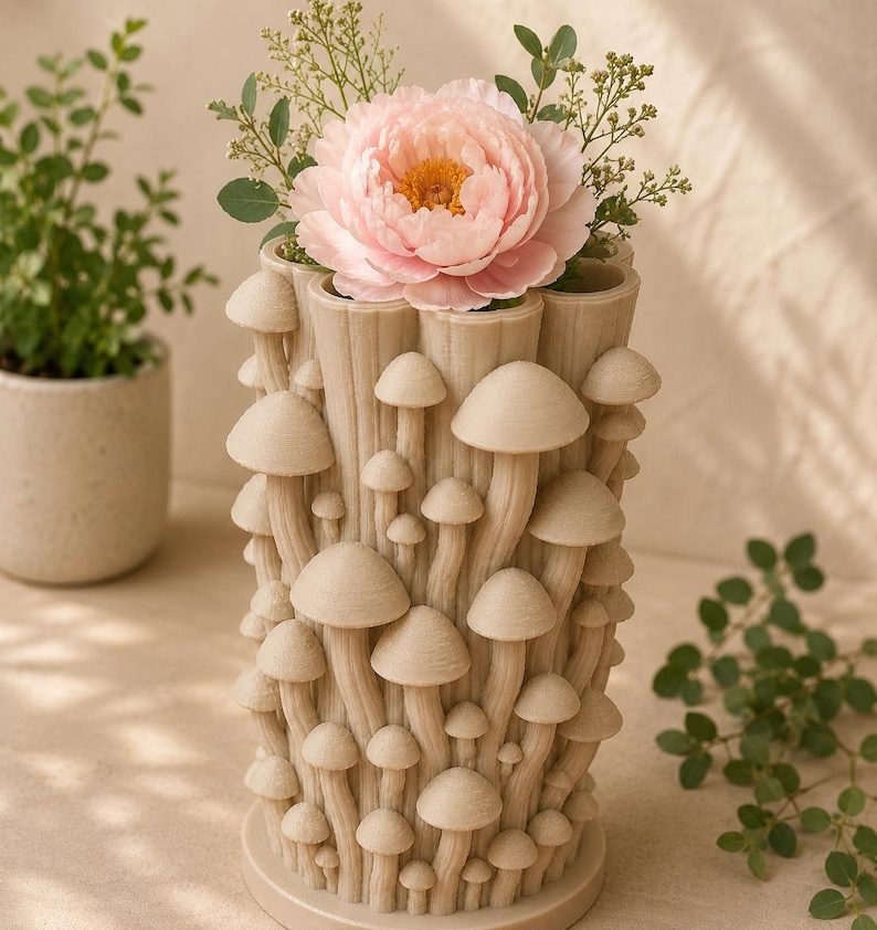 K&ouml;nnte beinhalten: Eine beige Vase mit Pilz-Motiv h&auml;lt eine gro&szlig;e rosa Pfingstrose und Gr&uuml;nzeug. Die zylindrische Vase hat eine strukturierte Oberfl&auml;che und ist mit Pilzformen in verschiedenen Gr&ouml;&szlig;en bedeckt. Die Gesamt&auml;sthetik ist skurril und von der Natur inspiriert.