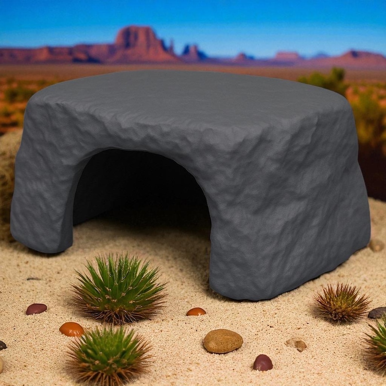 May include: A gray, rock-like reptile hideout with an arched opening. The hideout is textured to resemble stone and is placed on a sandy surface with small plants and pebbles. The background features a desert landscape.