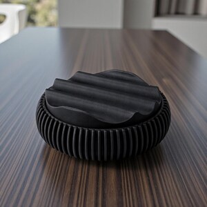 May include: A black ashtray with a ridged lid and a vertically ribbed base. The lid has a wavy design. The ashtray is made of a matte black material and sits on a dark wood surface.