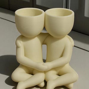 May include: A cream-coloured decorative planter featuring two figures embracing. Each figure has a rounded head with a hollowed-out top, suitable for holding small plants or flowers. The figures are seated with their legs crossed and arms around each other.