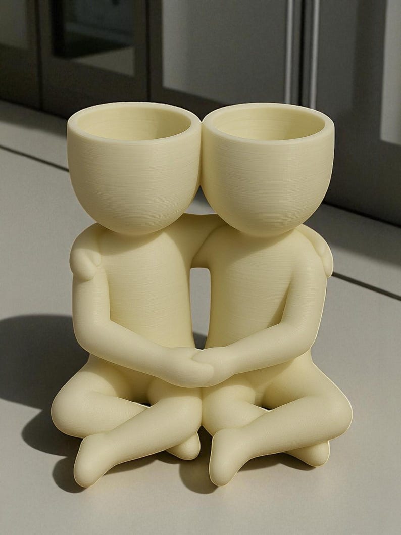 May include: A cream-coloured decorative planter featuring two figures embracing. Each figure has a rounded head with a hollowed-out top, suitable for holding small plants or flowers. The figures are seated with their legs crossed and arms around each other.