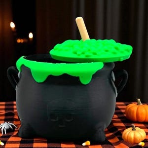 May include: A black cauldron with a green, bubbling substance and a wooden spoon. The cauldron has a skull design and is decorated with green slime. Small pumpkins and Halloween decorations are in the background.