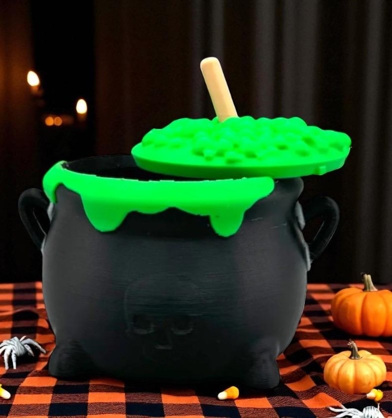 May include: A black cauldron with a green, bubbling substance and a wooden spoon. The cauldron has a skull design and is decorated with green slime. Small pumpkins and Halloween decorations are in the background.