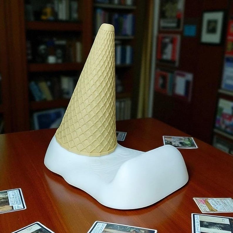May include: A whimsical ice cream cone phone stand. The stand features a light yellow cone atop a white, melted-ice-cream-shaped base. The cone has a textured waffle pattern. The stand is designed to hold a phone or other small device.