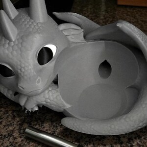 May include: A gray dragon-shaped sculpture with large eyes and wings. The dragon is holding a round object with a hole in the center. A silver metal tube with a black band is in the foreground.