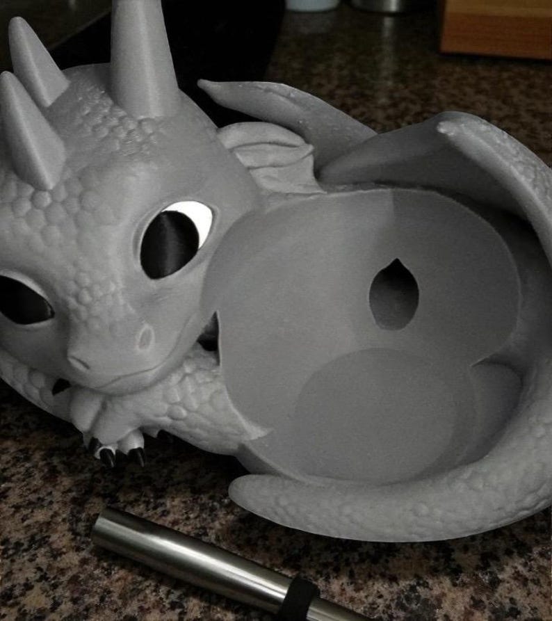 May include: A gray dragon-shaped sculpture with large eyes and wings. The dragon is holding a round object with a hole in the center. A silver metal tube with a black band is in the foreground.