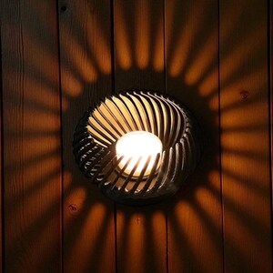 Pu&ograve; includere: Applique da parete decorativa con un design a spirale, che proietta un motivo di luce radiale e calda su una parete in legno. L'applique presenta una sorgente luminosa centrale, circondata da lamelle metalliche curve e scure.
