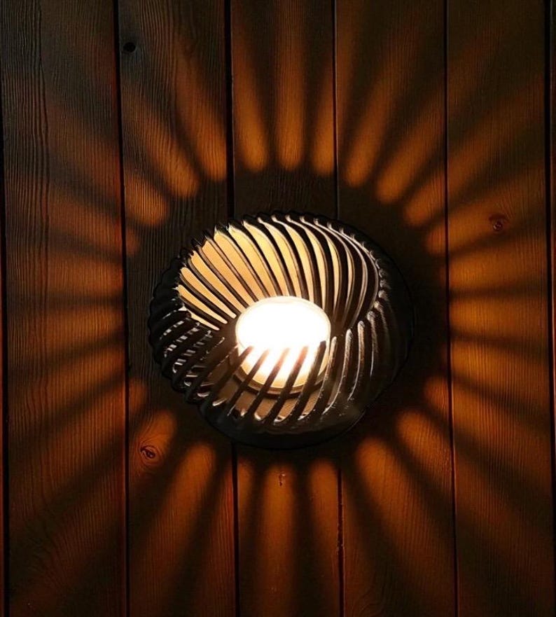 Pu&ograve; includere: Applique da parete decorativa con un design a spirale, che proietta un motivo di luce radiale e calda su una parete in legno. L'applique presenta una sorgente luminosa centrale, circondata da lamelle metalliche curve e scure.