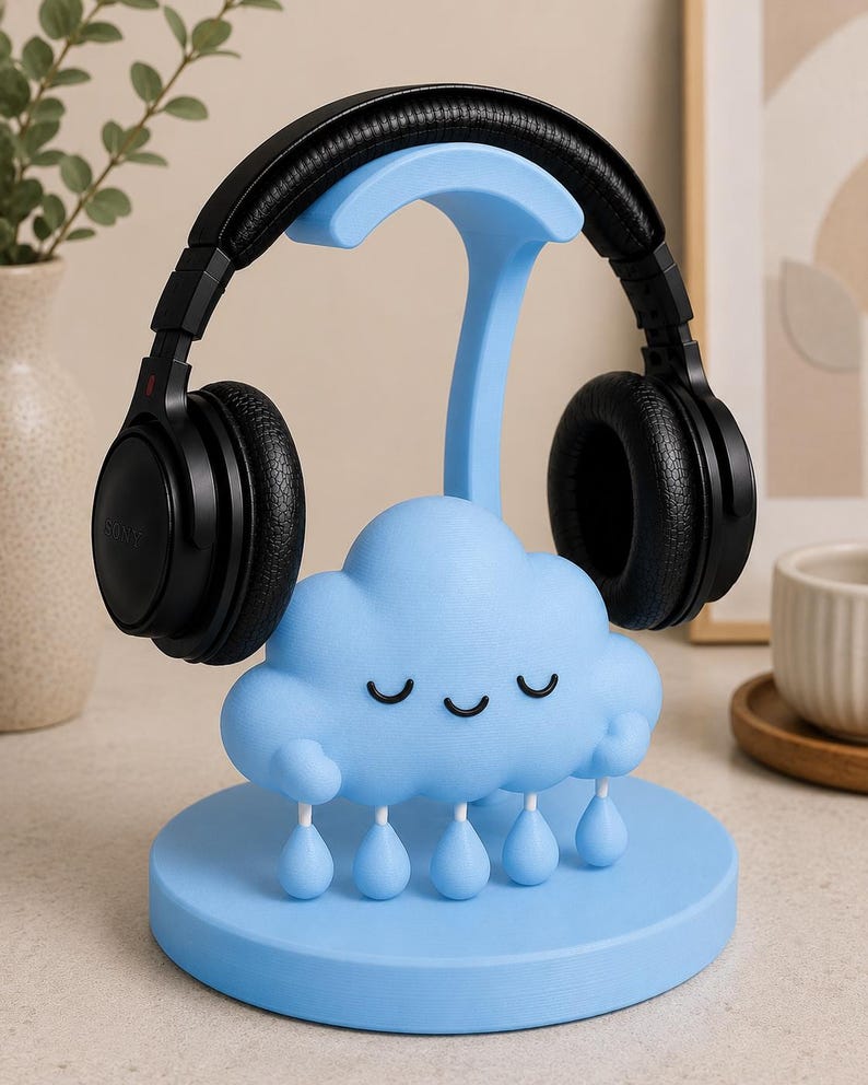 May include: A light blue headphone stand shaped like a cloud with raindrops, holding a pair of black headphones. The stand has a round base and a curved support for the headphones. The cloud has closed eyes.