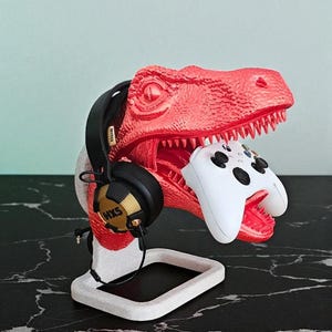 Könnte beinhalten: Ein roter Dinosaurier-Kopfhörer- und Controller-Ständer. Der Dinosaurier hat eine strukturierte Oberfläche und hält einen weißen Gamecontroller in seinem Maul. Schwarze Kopfhörer mit goldenen Akzenten ruhen auf dem Kopf des Dinosauriers. Der Ständer ist weiß.