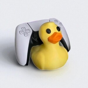 May include: A yellow rubber duck with black eyes and an orange beak is positioned in front of a white and black video game controller. The controller has a symmetrical design with a central touchpad and various buttons.