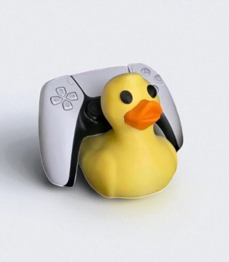 May include: A yellow rubber duck with black eyes and an orange beak is positioned in front of a white and black video game controller. The controller has a symmetrical design with a central touchpad and various buttons.