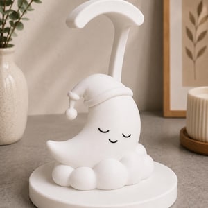 May include: A white night light shaped like a crescent moon wearing a sleeping cap, resting on a cloud-shaped base. The moon has closed eyes and a smiling expression. A curved handle extends above the moon. The night light is on a round base.