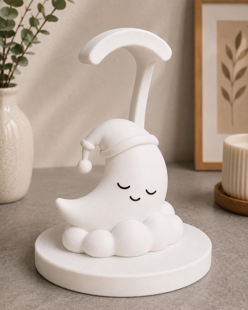 May include: A white night light shaped like a crescent moon wearing a sleeping cap, resting on a cloud-shaped base. The moon has closed eyes and a smiling expression. A curved handle extends above the moon. The night light is on a round base.