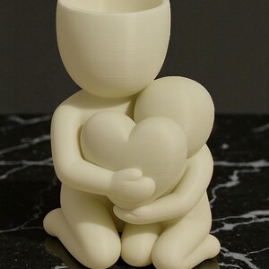 May include: A cream-colored figurine depicting two figures embracing a heart. The larger figure, with a cylindrical head, kneels, cradling a smaller figure and a heart-shaped object. The sculpture is made of a smooth, matte material.