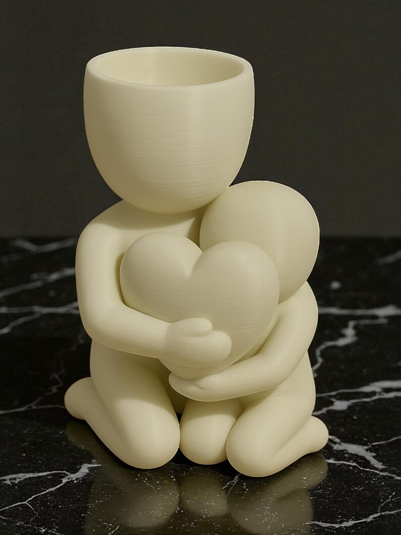 May include: A cream-colored figurine depicting two figures embracing a heart. The larger figure, with a cylindrical head, kneels, cradling a smaller figure and a heart-shaped object. The sculpture is made of a smooth, matte material.