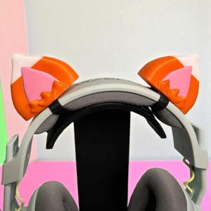 May include: A gray gaming headset with orange, white, and pink cat ear attachments. The headset is displayed on a black stand against a pink and white background. The ear attachments are shaped like cat ears.