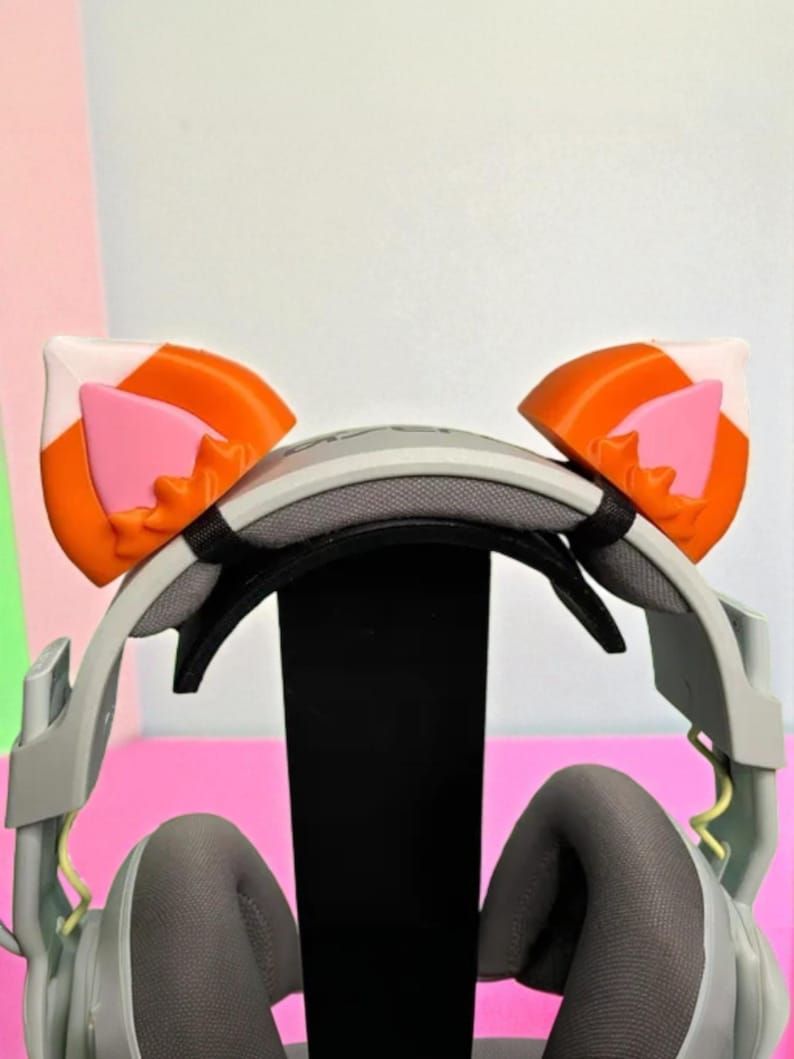 May include: A gray gaming headset with orange, white, and pink cat ear attachments. The headset is displayed on a black stand against a pink and white background. The ear attachments are shaped like cat ears.