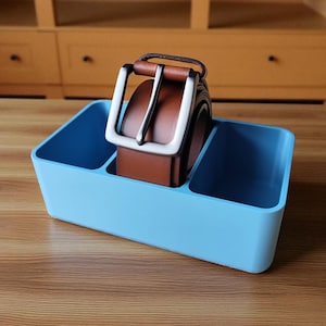 Puede incluir: Un organizador rectangular azul claro con tres compartimentos, que contiene un cintur&oacute;n de cuero marr&oacute;n con una hebilla plateada. El organizador est&aacute; sobre una superficie de madera, con un fondo borroso de un mueble de madera.