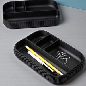 May include: Two black desk organizers with multiple compartments. One organizer is empty, while the other contains a yellow pencil, a white pencil, a black pen, and silver paper clips. The organizers have a ribbed exterior.