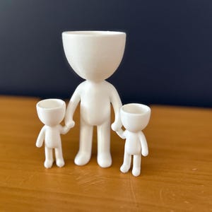 May include: A white ceramic family figurine set. The set includes a large figure holding the hands of two smaller figures. Each figure has a rounded head and a simple, minimalist design. The figures are displayed on a wooden surface.