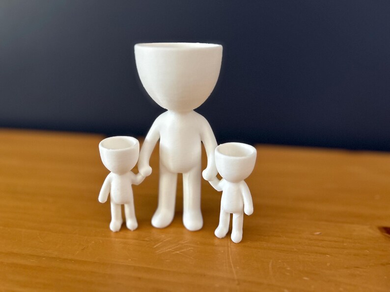 May include: A white ceramic family figurine set. The set includes a large figure holding the hands of two smaller figures. Each figure has a rounded head and a simple, minimalist design. The figures are displayed on a wooden surface.