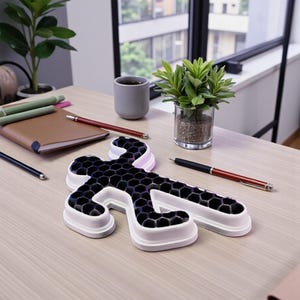 Op de afbeelding: Een ijsvorm in de vorm van een persoon met een witte rand en een zwart honingraatpatroon ligt op een licht houten bureau. Pennen, een notitieboek en kleine potplanten staan ook op het bureau. Een kopje koffie staat bij een raam.