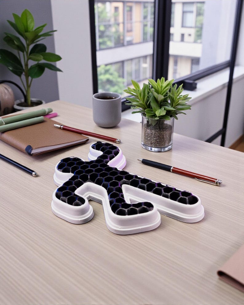 Op de afbeelding: Een ijsvorm in de vorm van een persoon met een witte rand en een zwart honingraatpatroon ligt op een licht houten bureau. Pennen, een notitieboek en kleine potplanten staan ook op het bureau. Een kopje koffie staat bij een raam.