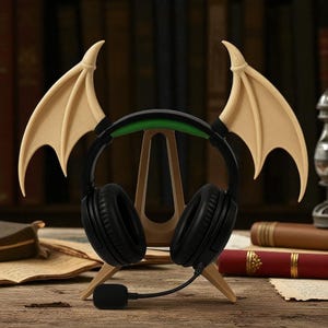 May include: Black gaming headphones with a green headband and attached microphone, displayed on a wooden stand. The stand is adorned with beige bat wings on either side, creating a unique aesthetic. The setup is on a wooden surface.
