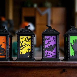 May include: Four black Halloween lanterns with different colored backgrounds and spooky cutouts. The lanterns feature orange with a pumpkin, yellow with a graveyard, purple with a bat, and green with a zombie hand. The lanterns are on a wooden surface.