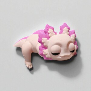 May include: A light pink axolotl figurine with bright pink gills and accents. The axolotl has closed eyes and a relaxed expression. The figurine is lying down on a light gray surface.