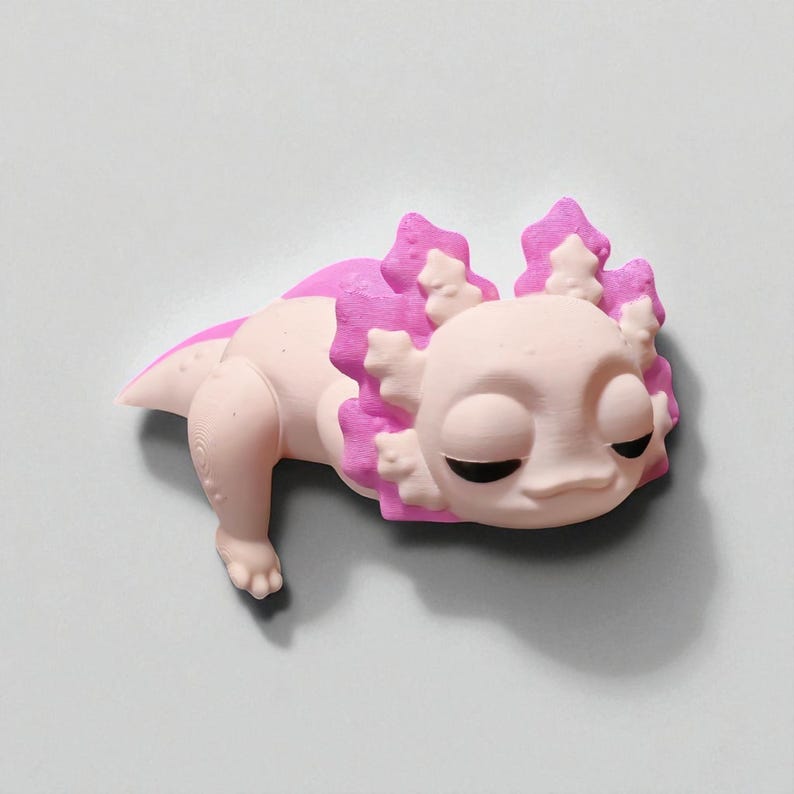 May include: A light pink axolotl figurine with bright pink gills and accents. The axolotl has closed eyes and a relaxed expression. The figurine is lying down on a light gray surface.