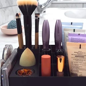 May include: A black makeup organizer filled with various cosmetic items. Includes makeup brushes, mascara tubes, lipsticks, a makeup sponge, and tubes of skincare products. The brushes have black handles and beige bristles. The organizer sits on a white countertop.