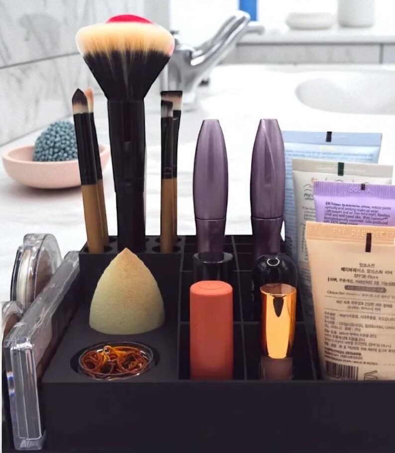 May include: A black makeup organizer filled with various cosmetic items. Includes makeup brushes, mascara tubes, lipsticks, a makeup sponge, and tubes of skincare products. The brushes have black handles and beige bristles. The organizer sits on a white countertop.